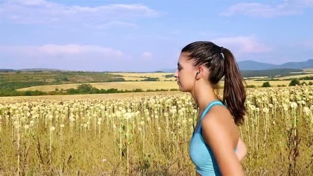 Cámara de seguimiento de mujer chica con auriculares corriendo corriendo en el campo de girasol, cámara lenta — Vídeos de Stock