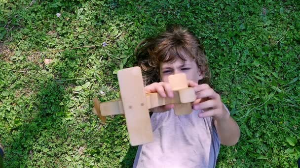 Boy playing with wooden toy airplane lying on the green grass, top view — Stockvideo