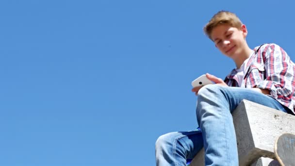 Nízký úhel pohledu dospívající chlapec psaní SMS, SMS na smartphone proti modré obloze — Stock video