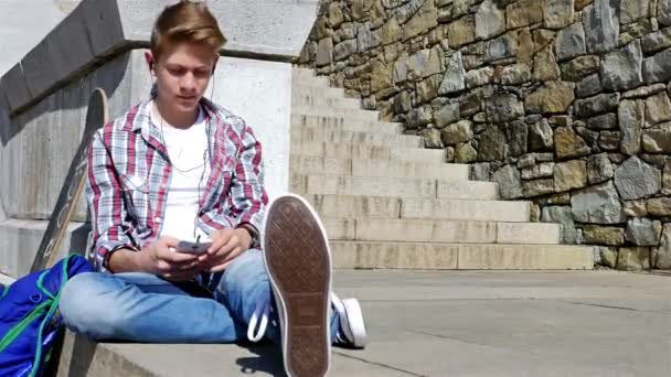 Tiener jongen met skateboard en rugzak luisteren naar muziek op smartphone zittend op de trap — Stockvideo