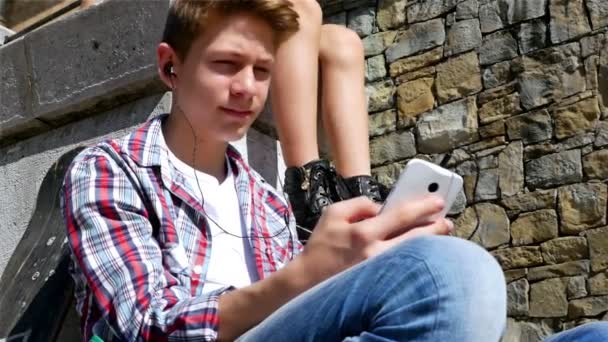 Мальчик-подросток со скейтбордом и рюкзаком слушает музыку на смартфоне — стоковое видео