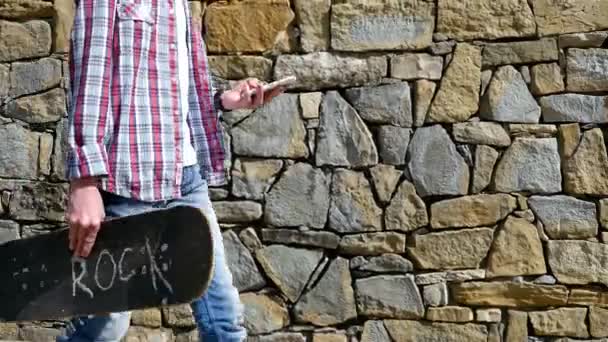 Teenager boy with skateboard walking using smartphone in the city — Stock Video