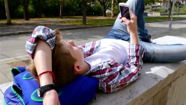 Adolescente chico escribiendo sms, mensajes de texto en el teléfono inteligente que se encuentra en el parque — Vídeo de stock