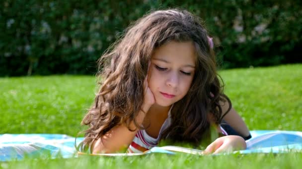 Petite fille mignonne aux cheveux bouclés lit un livre dans le jardin — Video