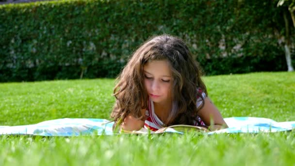 Cute little girl with curly hair is reading a book in the garden — Stock Video