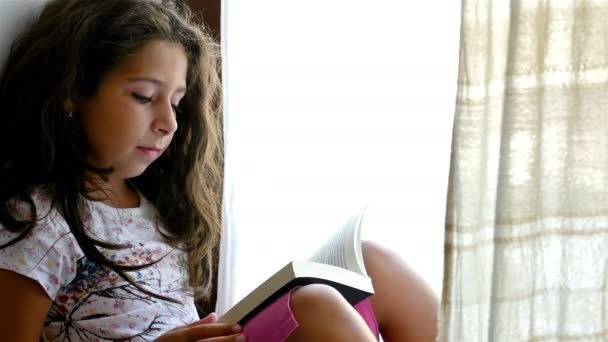 Menina bonito sentado no peitoril da janela de uma casa velha, lendo livro para seu brinquedo urso — Vídeo de Stock