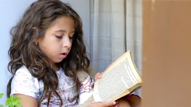 Menina bonito sentado no peitoril da janela de uma casa velha, lendo livro para seu brinquedo urso — Vídeo de Stock