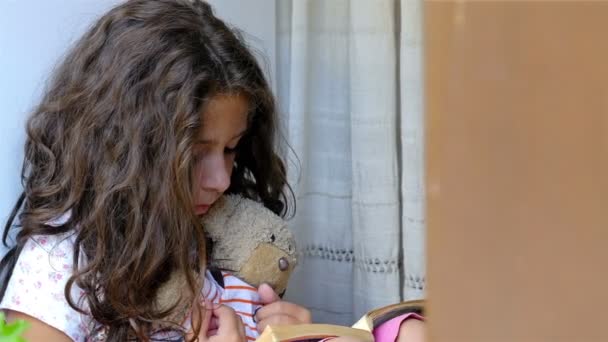 Милая маленькая девочка, сидящая на подоконнике старого дома и читающая книгу своей медвежьей игрушке — стоковое видео