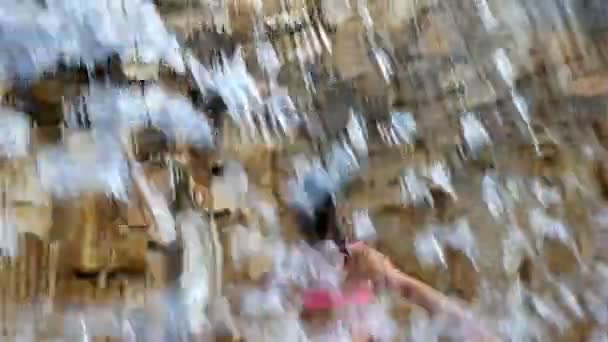 Nettes kleines Mädchen im Badeanzug hat Spaß unter fallendem Wasser vor einer Steinmauer — Stockvideo