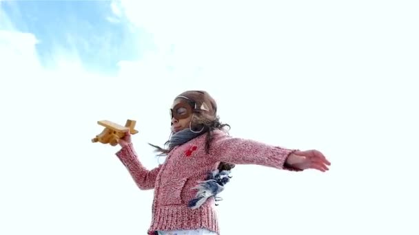 Niña con sombrero de piloto jugando con avión de madera contra el cielo azul, cámara lenta — Vídeos de Stock