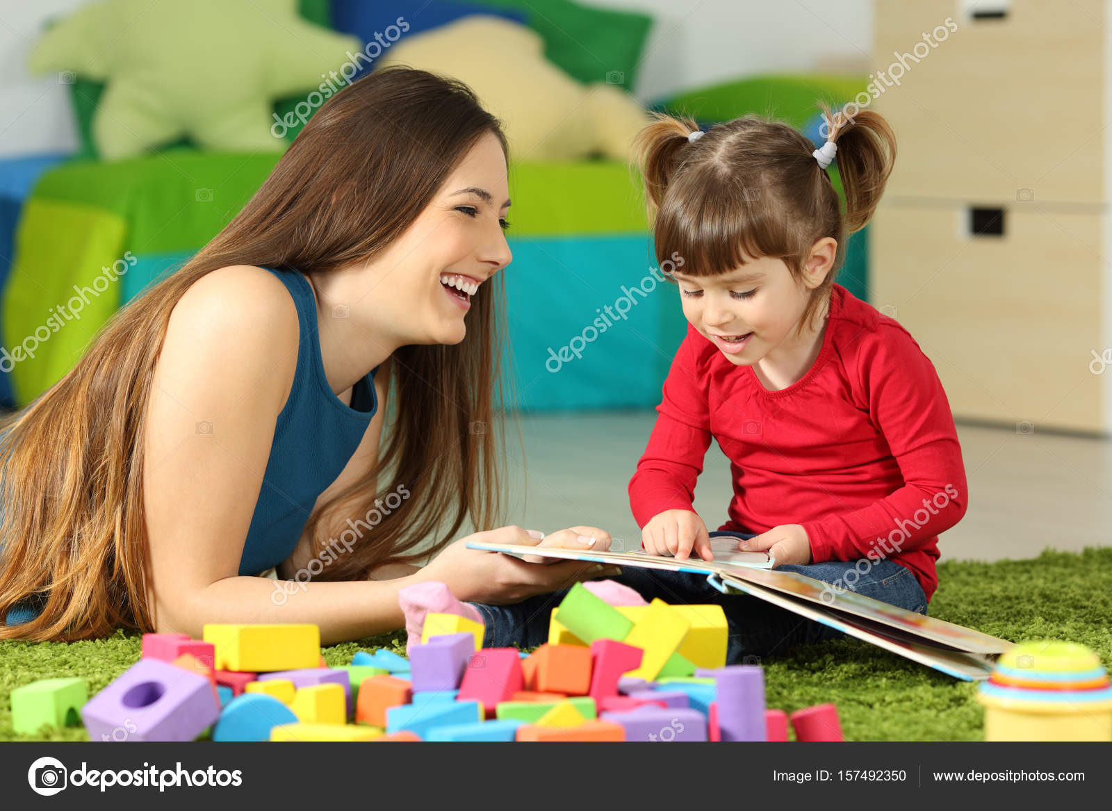 Madre e bambino che gioca insieme ad un libro sdraiato sul pavimento della camera da letto a casa con uno sfondo colorato — Foto di AntonioGuillemF