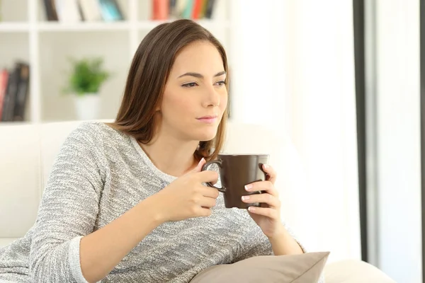 Chica pensativa sosteniendo una taza de café en un sofá en casa — Foto de Stock