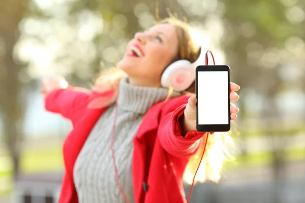 Chica alegre escuchando música y mostrando la pantalla del teléfono en invierno — Foto de Stock