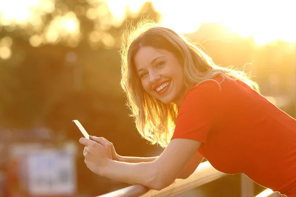 Szczęśliwa dziewczyna patrzy na Ciebie z inteligentny telefon o zachodzie słońca — Zdjęcie stockowe
