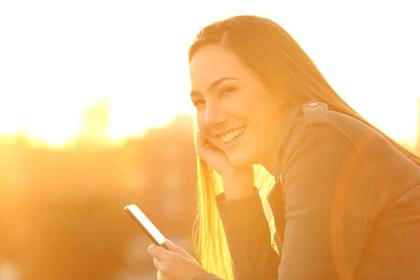 Flicka som håller en smartphone som tittar på dig i solnedgången — Stockfoto