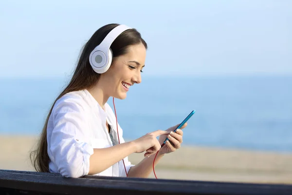 Mujer escuchando música sentada en un banco en la playa — Foto de Stock