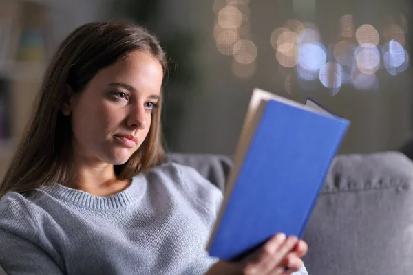 Mujer seria leyendo un libro de papel en un sofá en la noche — Foto de Stock