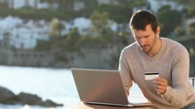 Ciddi bir adam sahilde bir balkonda dizüstü bilgisayar ve kredi kartıyla ödeme yapıyor.