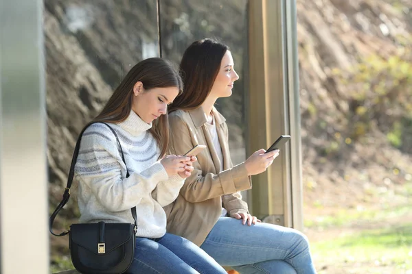 Дві жінки чекають на автобусній зупинці, перевіряючи телефон — стокове фото