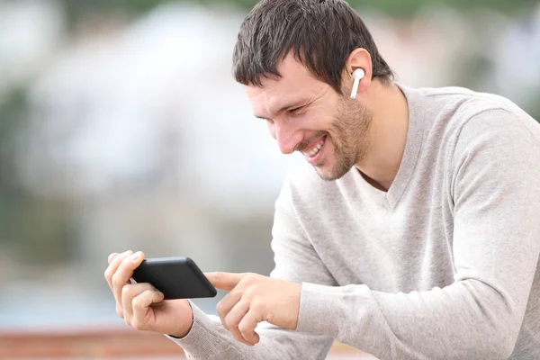 Onnellinen mies katselee ja kuuntelee mediaa puhelimella ja kuulokkeilla — kuvapankkivalokuva