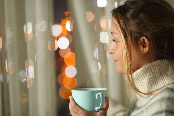 Mujer sosteniendo taza de café mirando por una ventana por la noche —  Fotos de Stock
