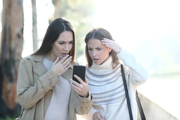 Dwie zmartwione kobiety sprawdzające wiadomości telefoniczne w parku — Zdjęcie stockowe