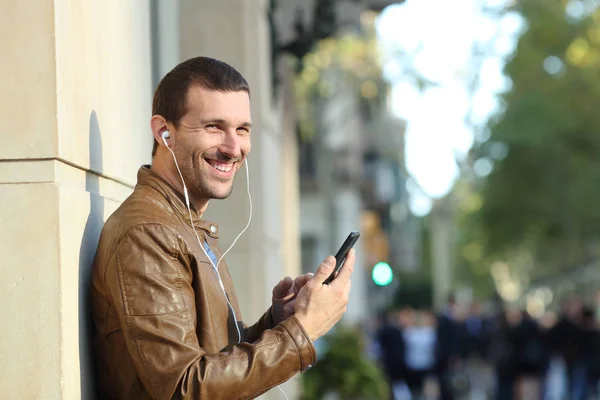 Happy man wearing earphones holding phone looks at you — 스톡 사진