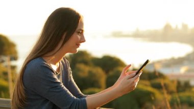 Gün batımında balkonda akıllı bir telefon kullanan rahat bir kadın.