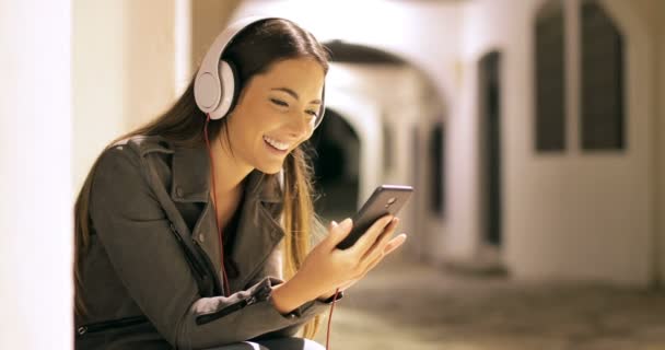 夜の街に座っているスマートフォンから音楽を聴く幸せな女の子 — ストック動画