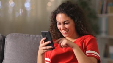 Evdeki oturma odasındaki koltukta oturan kırmızı elbiseli, akıllı telefonları kontrol eden ciddi bir Arap kızı. 