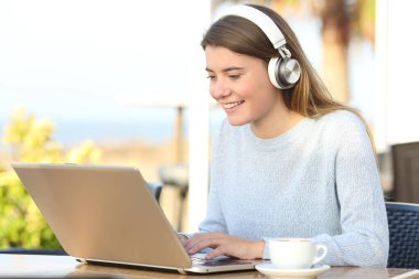 Kafe terasında oturan kulaklıklı ve dizüstü bilgisayarlı mutlu öğrenci e-öğrenimi