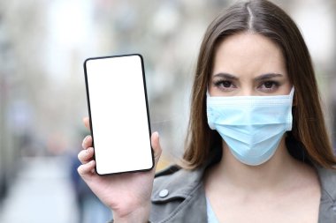 Worried woman wearing protective mask showing smart phone blank screen in the street