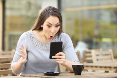 Amazed girl looking surprised at her smart phone on a cafe terrace