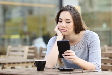 Bored girl looking disappointed at her smart phone on a cafe terrace