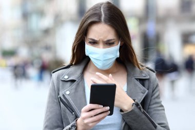 Front view portrait of a scared girl wearing protective mask avoiding contagion reading news on her smart phone on a city street