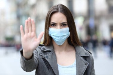Front view portrait of a serious girl doing stop sign gesture with protective mask on city street