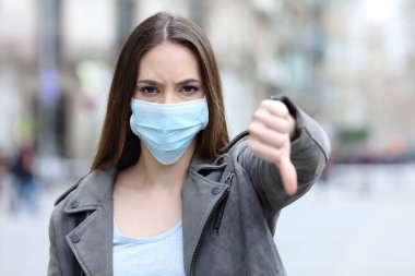 Front view portrait of serious girl doing thumbs down gesture with protective mask on city street