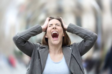 Front view portrait of a stressed woman screaming desperate suffering anxiety attack grabbing her head on city street
