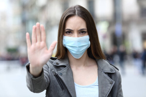 Портрет серьезной девушки, делающей знак "стоп" в защитной маске на городской улице
