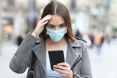 Koruyucu maskeli endişeli bir kadının portresi şehrin caddesinde akıllı telefondan haberleri kontrol ediyor.