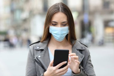 Şehir caddesinde akıllı telefonunu kontrol eden, koruyucu maskeli, ciddi bir genç kızın portresi.