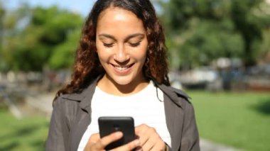 Güneşli bir günde parkta akıllı telefon kullanan mutlu bir melez kadının ön görüntüsü.