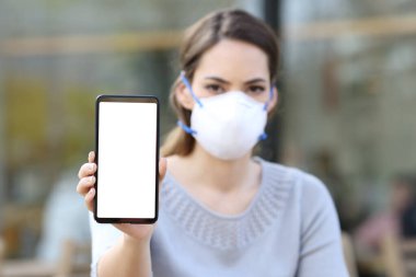 Koruyucu maskeli ciddi bir kadının portresi kafeteryadaki boş ekranlı akıllı telefonu gösteriyor.
