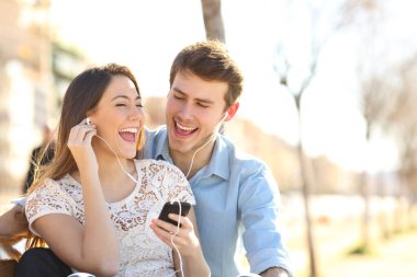 Mutlu çift birlikte şarkı söylüyor, akıllı telefondan müzik dinliyor, kulaklıklarını paylaşıyor, parkta bir bankta oturuyor.