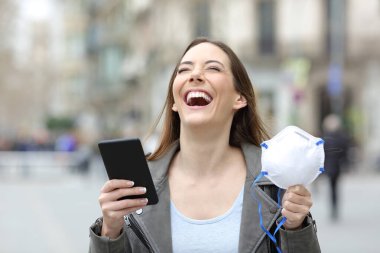 Front view portrait of a excited woman holding smart phone and protective mask celebrating good news in a city street clipart