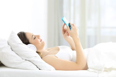 Rahat bir kızın yan görünüm portresi müzik dinleyen akıllı telefondan şarkı seçen evde bir yatakta uzanan