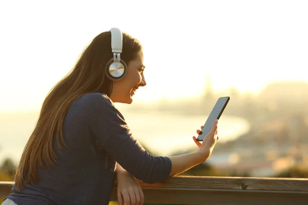 幸せな女の子のプロフィール日没時に公園にヘッドフォンでスマートフォンで音楽を聴く — ストック写真