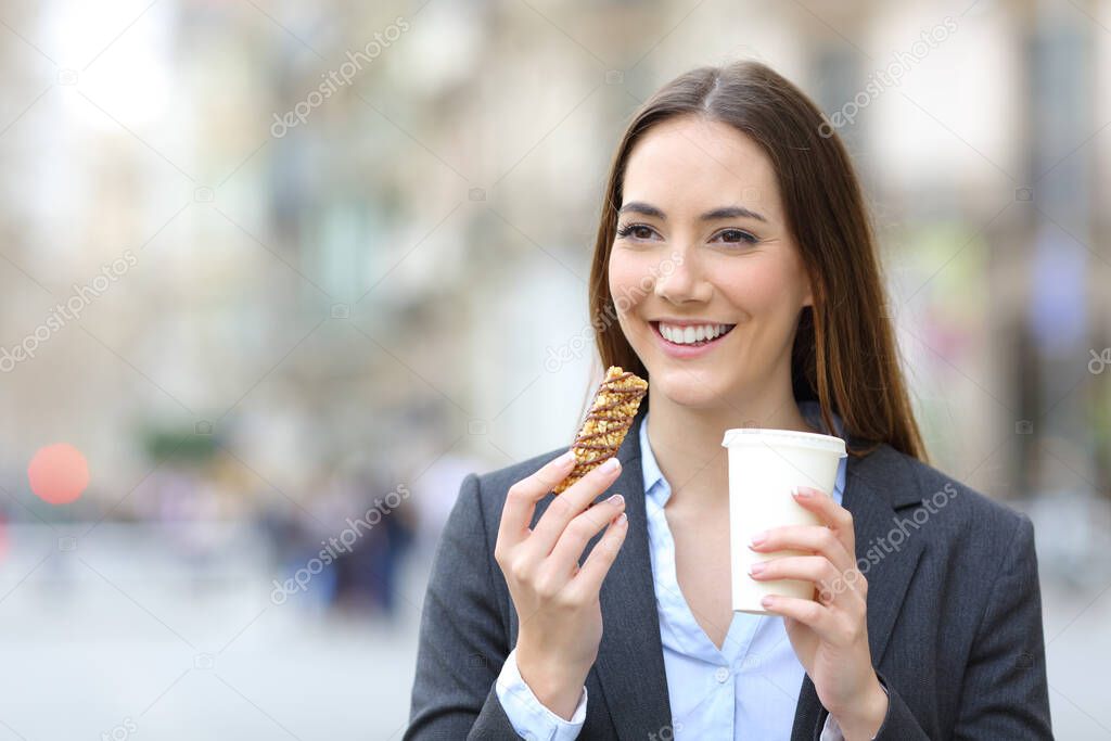 Front view portrait of a happy executive woman holding cereal snack bar and takeaway coffee in the street
