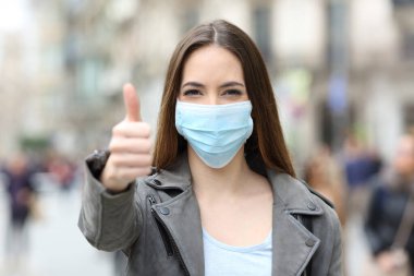 Koruyucu maskeli mutlu bir kadının ön manzara portresi. Sokakta baş parmak işareti yapıyor.