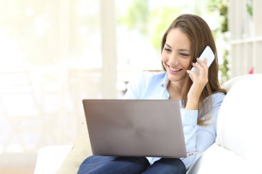 Bilgisayarla çalışan mutlu kız akıllı telefondan arıyor. Evdeki koltukta oturuyor.
